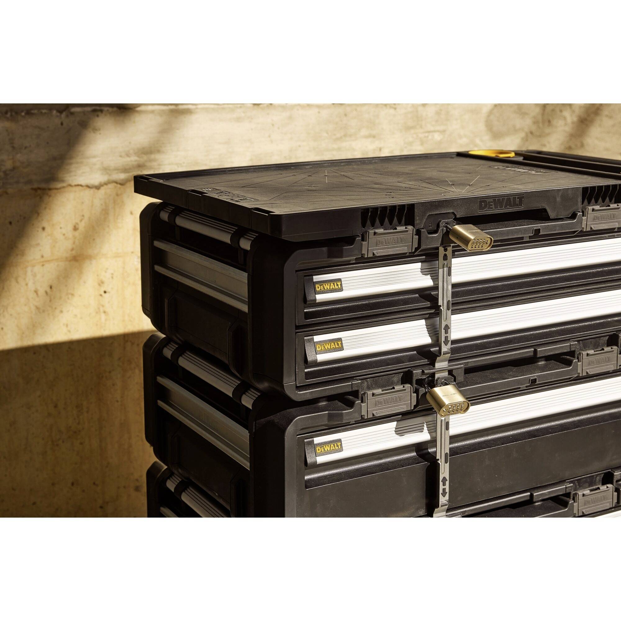 Svarta förvaringslådor av plast med metallås, staplade framför en betongvägg i solljuset.