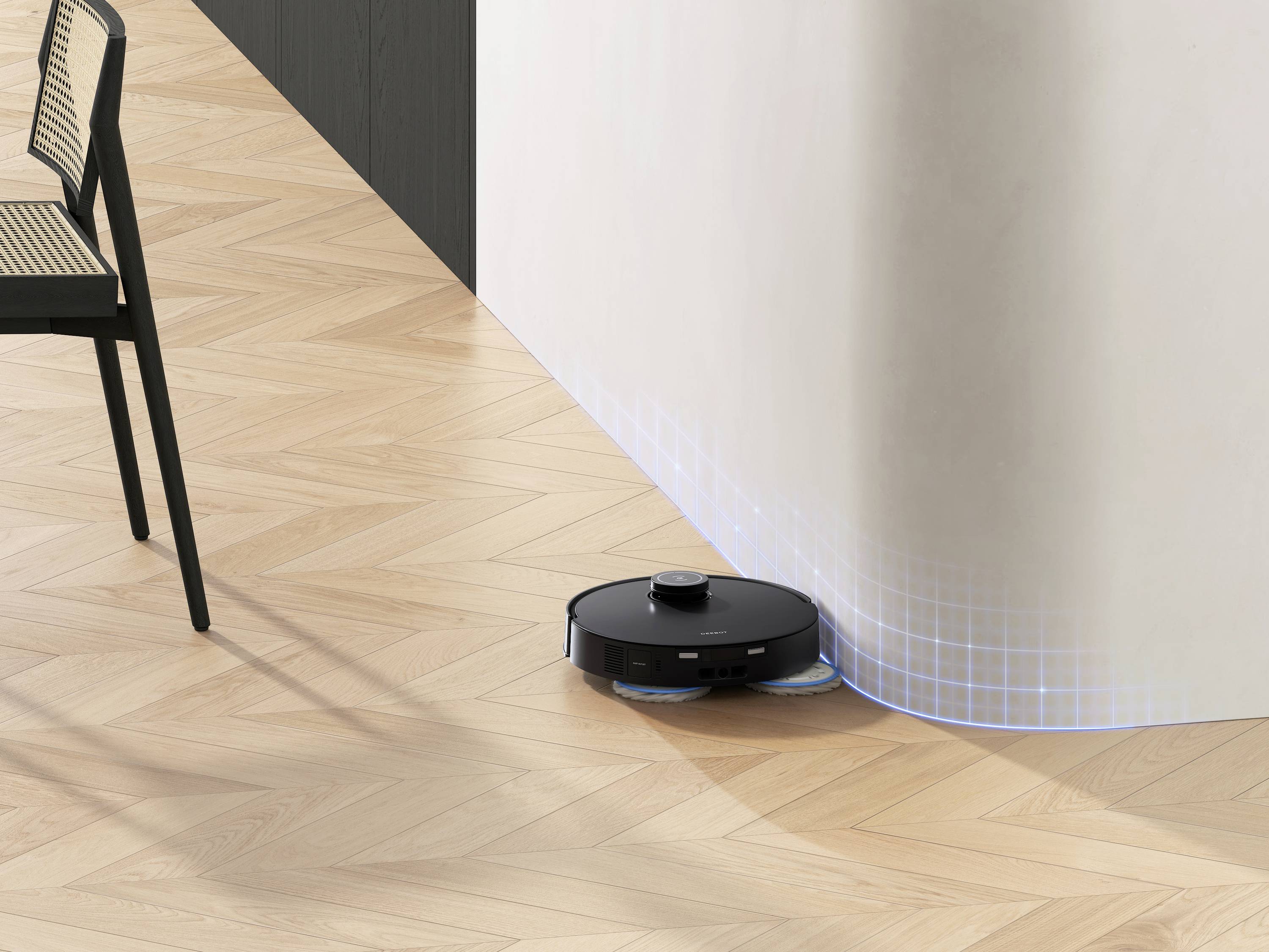 En svart robotdammsugare rengör trägolvet bredvid en böjd vägg. En stol står till vänster i bilden.