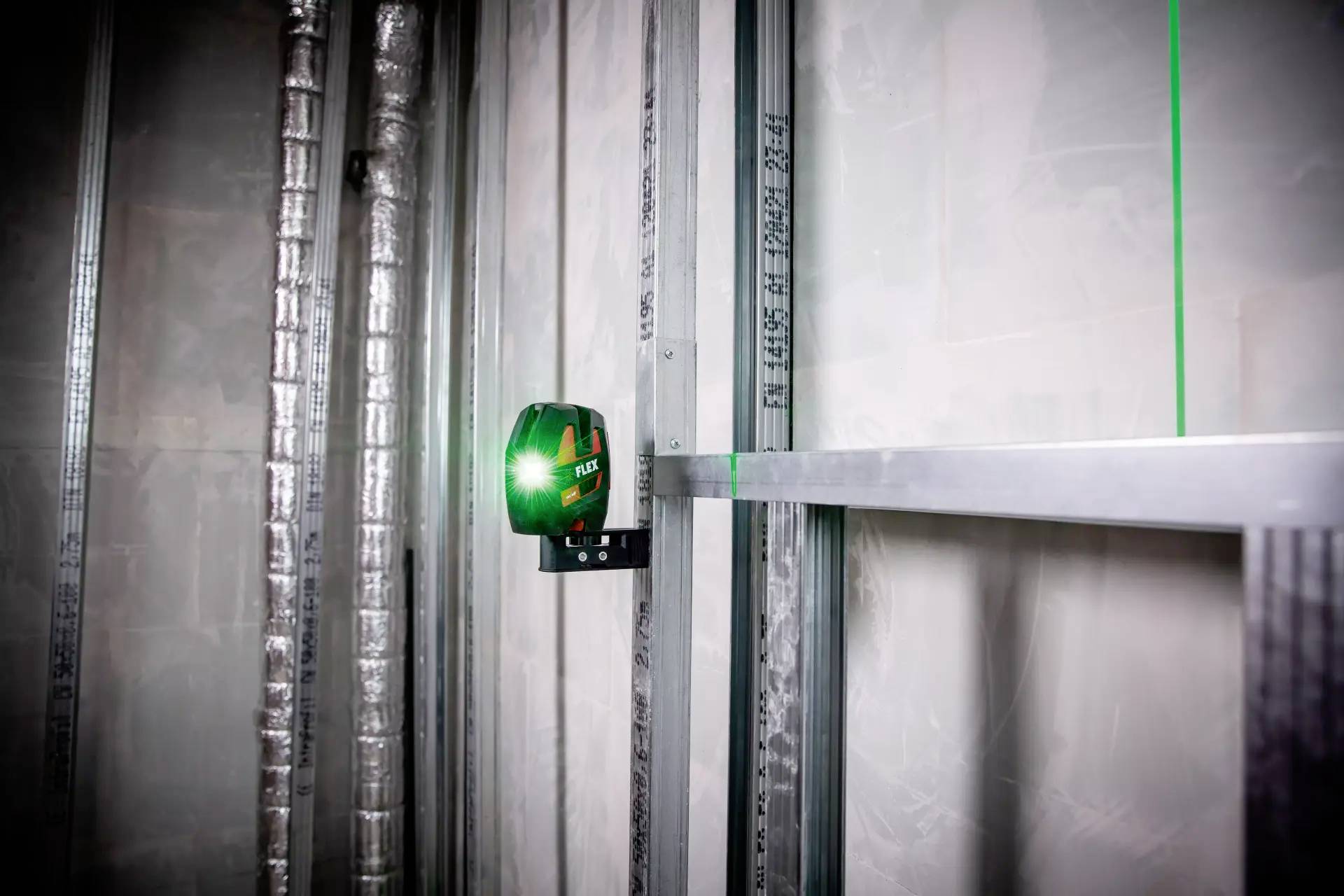 Elektronisk enhet projicerar ett grönt lasernivåplan på väggen i ett rum med metallram och isolering för att stödja byggnadsarbeten.