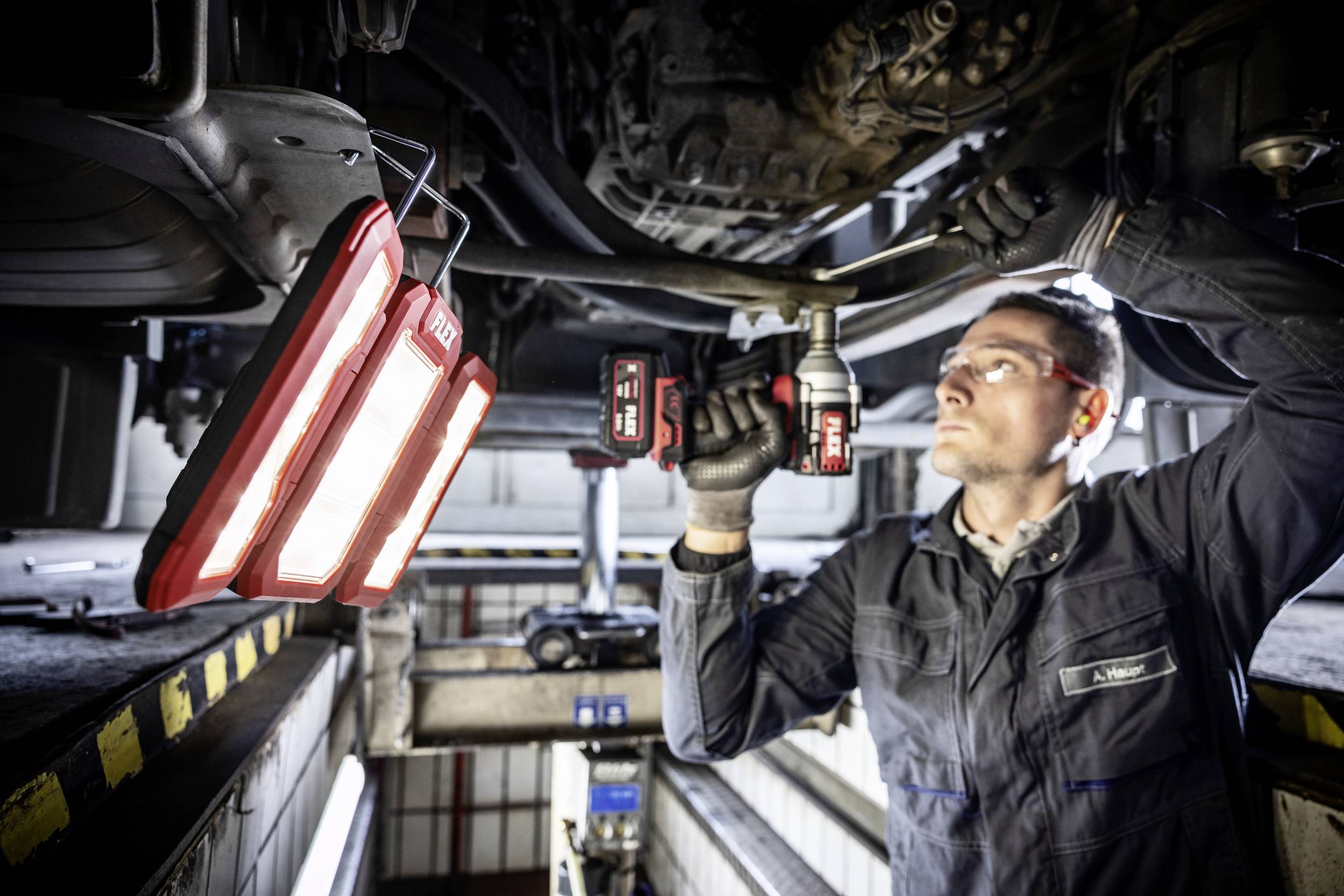 En mekaniker arbetar under en upplyft bil, håller ett elverktyg och en lampa lyser upp arbetsplatsen.