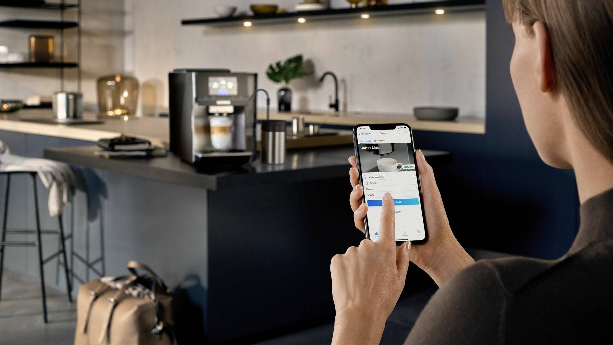 En person styr en kaffebryggare i ett modernt kök via en smartphoneapp. Maskinen tillagar en kopp kaffe.