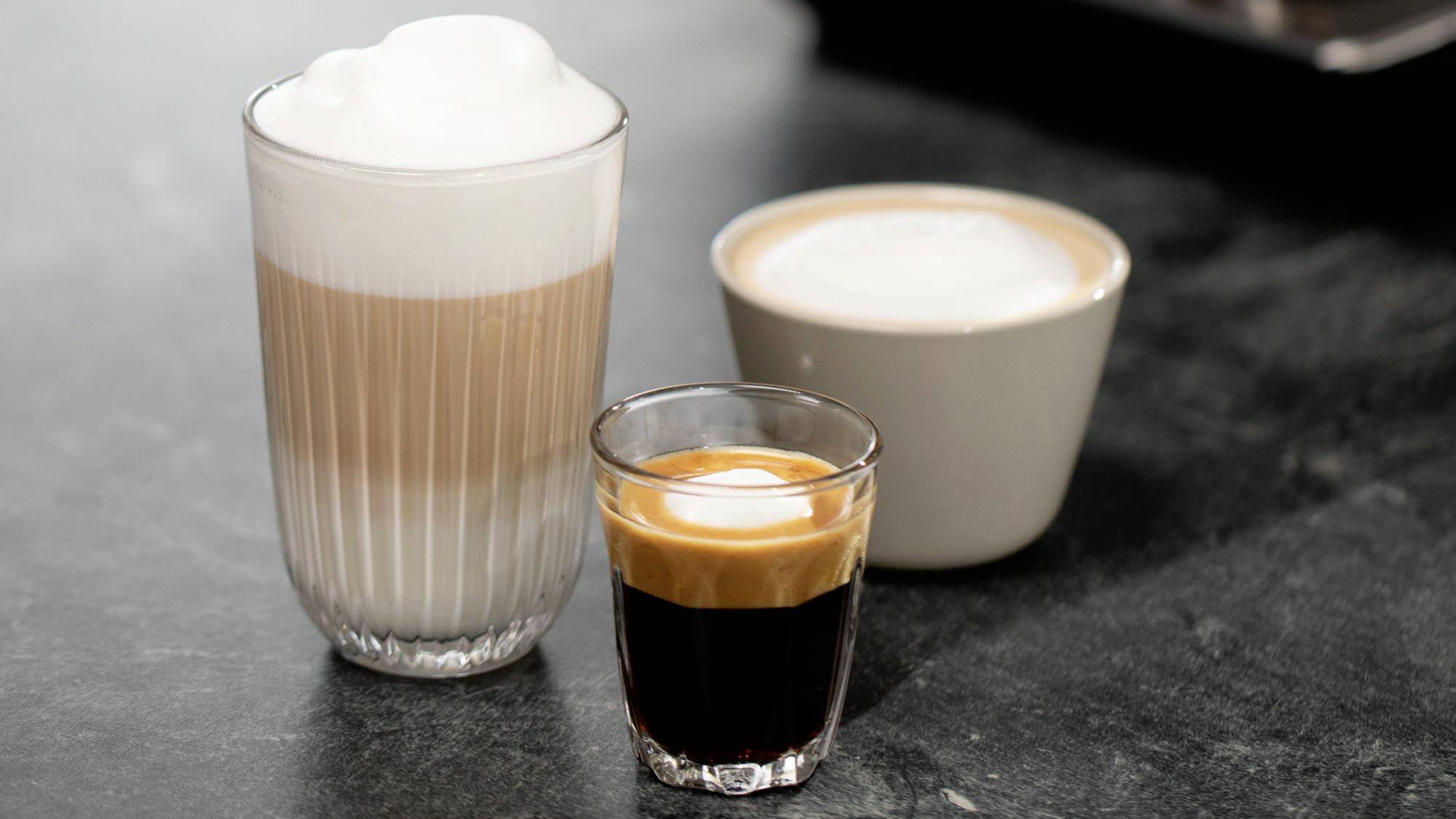 Tre olika kaffedrycker: Ett glas Latte Macchiato, en kopp Cappuccino och en Espresso i ett litet glas på ett mörkt bord.