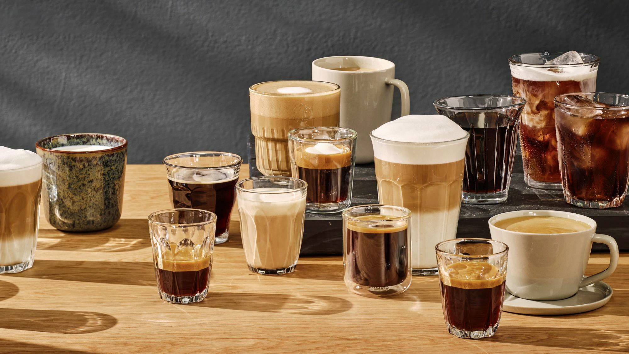 Ett urval av olika kaffessorter i glas och koppar, inklusive espresso, cappuccino, latte och iskaffe, på ett träbord.