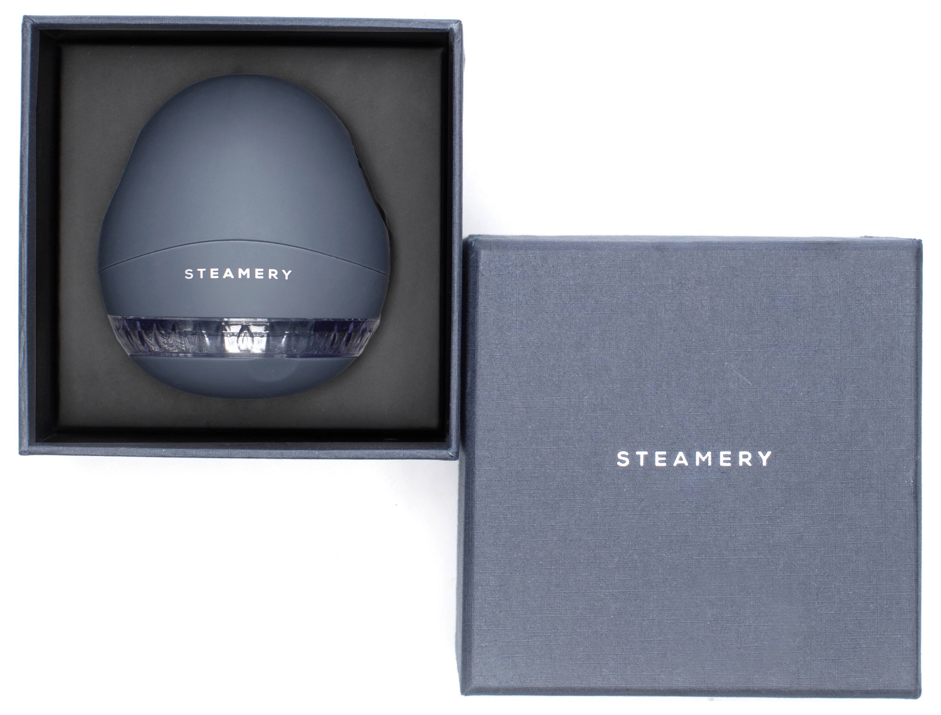 En blå apparat från märket 'Steamery' i en öppnad låda. Lådan bredvid visar 'Steamery'-logotypen på locket.