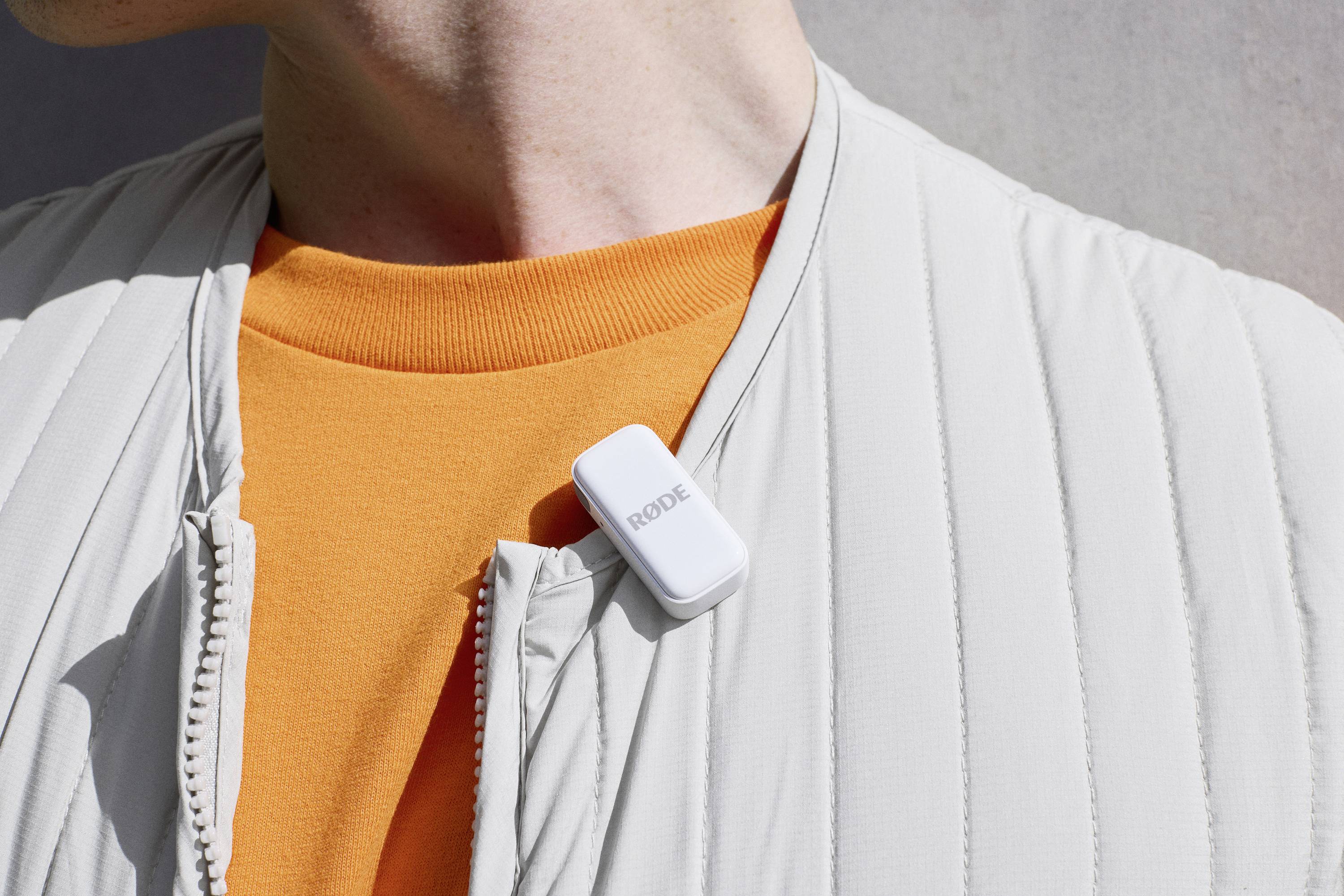 En vit trådlös mikrofon är fäst vid en ljusgrå jacka framför en orange tröja.