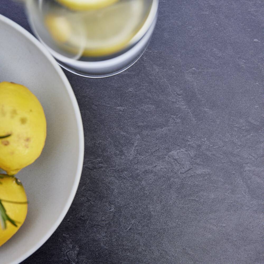 Två citroner i en skål bredvid ett glas med citronskivor på ett mörkt, strukturerat underlag.