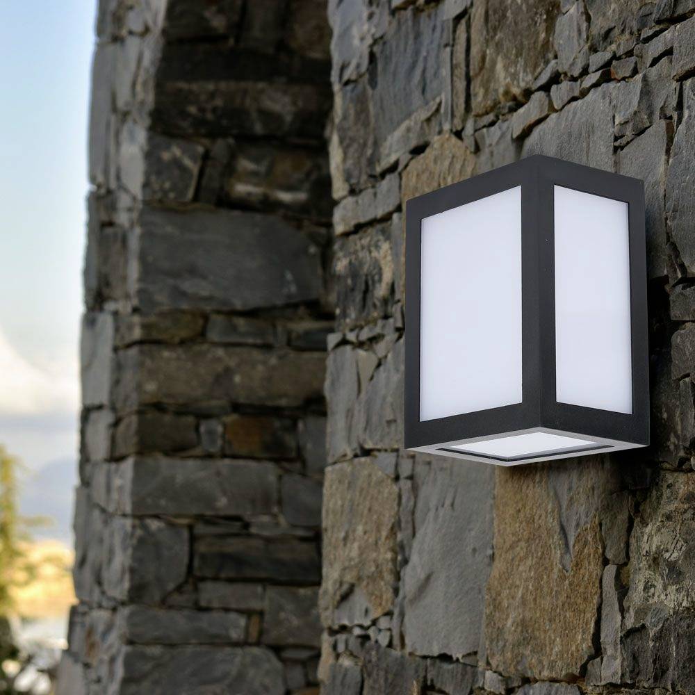 Vägglampa av svart metall och glas monterad på en stenmur, med ett mjukt sken. Omgivningen visar stenstruktur.