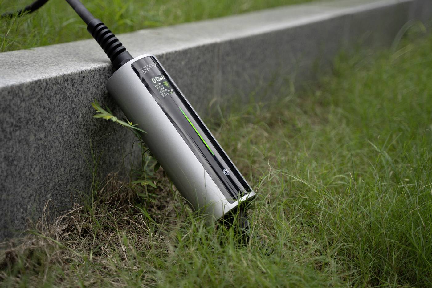 En laddningskabel ligger i gräset, ansluten till en elektriskt driven enhet. Enheten har en tydlig laddningsindikator.