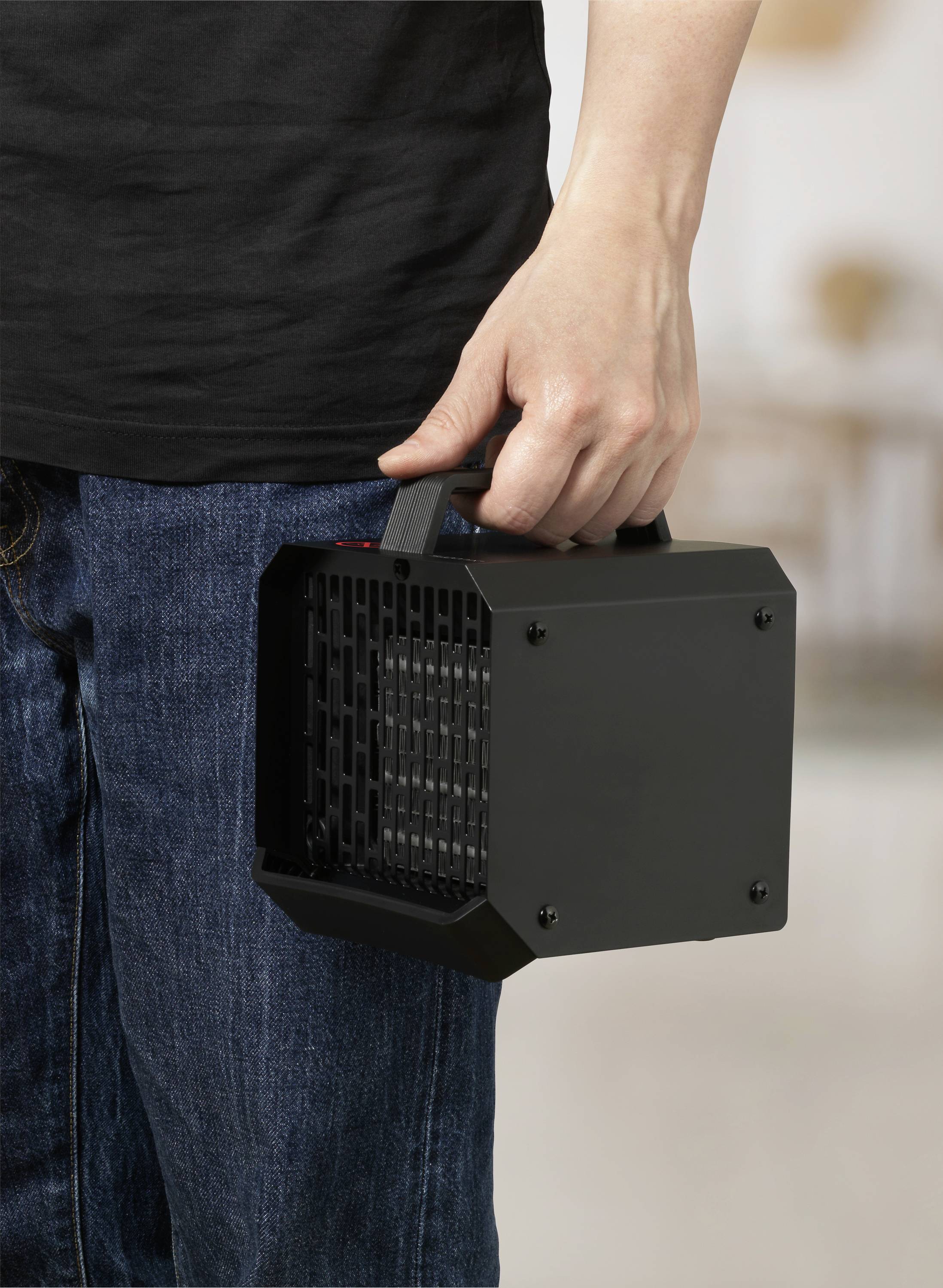En person håller en bärbar, svart värmefläkt med ett handtag. Personen bär jeans och en svart överdel. Värmefläkten har ett gallerdésign.
