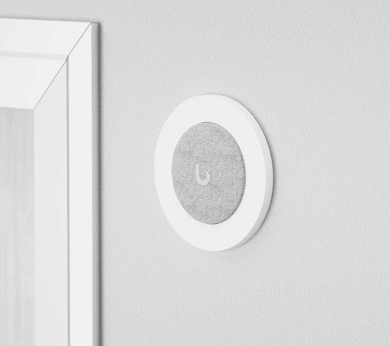 En rund, väggmonterad smart hemapparat bredvid en dörrpost, med ett diskret grått tygöverdrag i mitten.