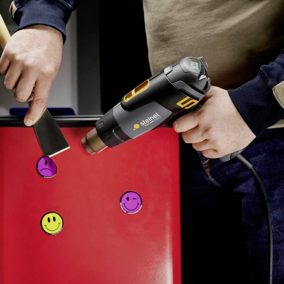 En person håller ett värmepistol över ett rött skåp med färgglada klistermärken av smiley-ansikten för att ta bort dem.