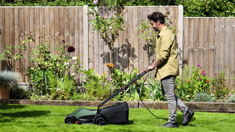 En man klipper gräs i en trädgård med en elektrisk gräsklippare. I bakgrunden finns ett trästaket och blommande växter.