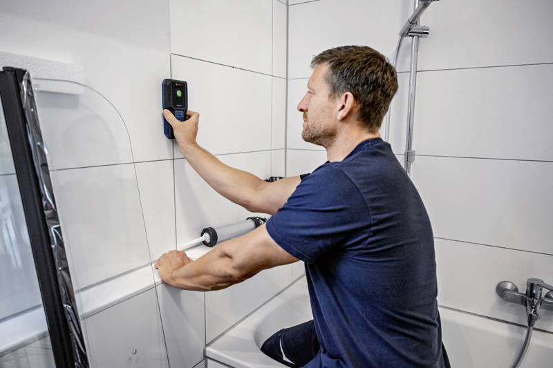 En man installerar en elektrisk apparat ovanför ett badkar. Badrummet har vita kakel och ett stödhandtag är monterat på väggen.