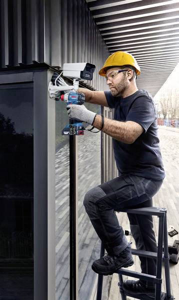 En arbetare med skyddshjälm installerar en övervakningskamera på en modern byggnad. Han står på en stege och använder verktyg.