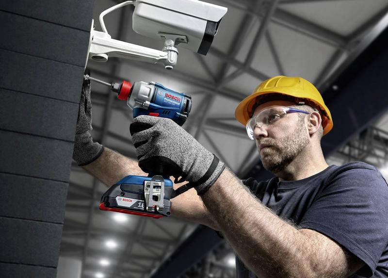 En byggnadsarbetare med skyddshjälm och handskar monterar en övervakningskamera på en vägg i en industriell miljö.