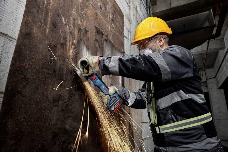 En byggnadsarbetare med skyddshjälm och skyddsglasögon klipper en metallplatta med en vinkelslip, gnistor flyger åt alla håll.