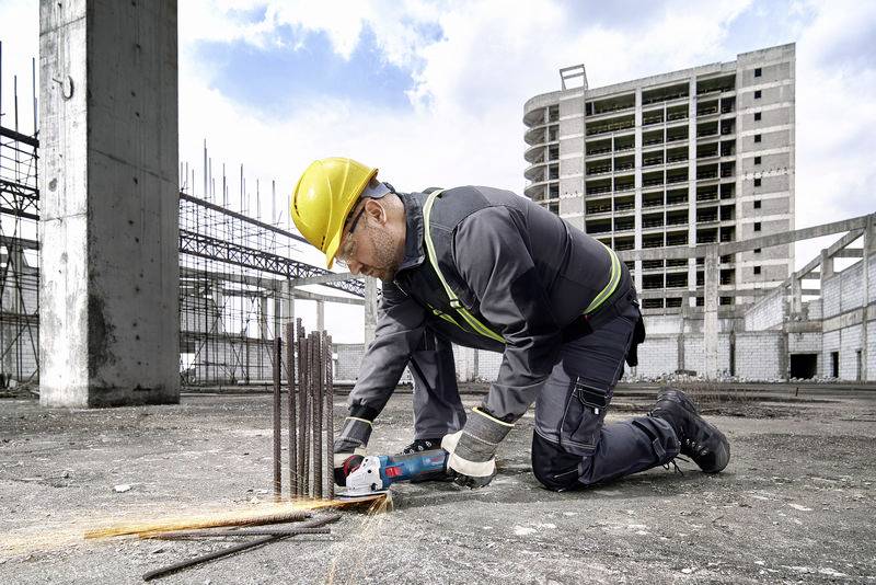 Byggarbetare på en byggarbetsplats, knäböjer och klipper metallstänger med en vinkelslip. Byggnadsställning och en ofärdig byggnad syns i bakgrunden.