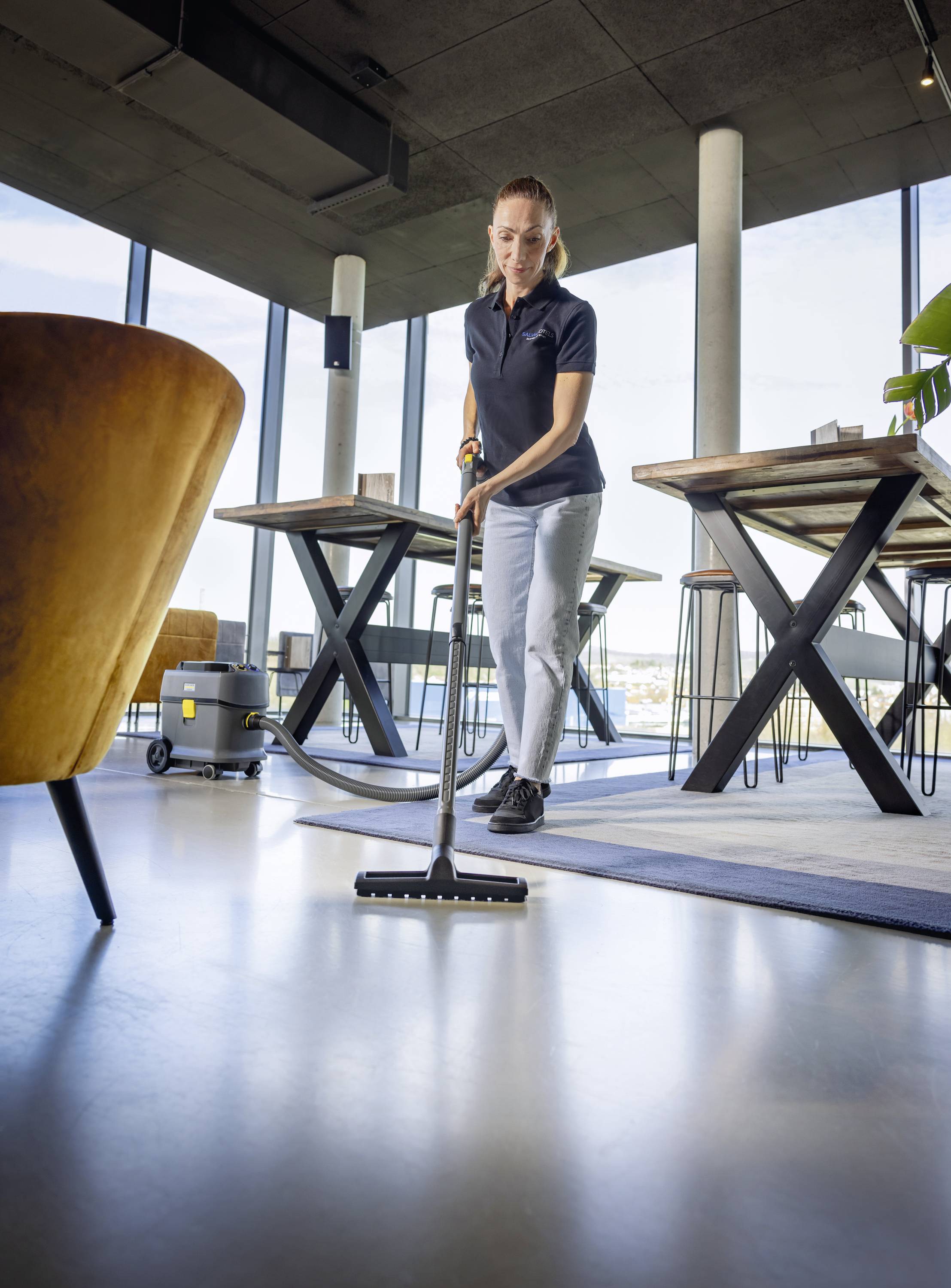 En person städar golvet i ett modernt, ljust rum med en dammsugare. I bakgrunden syns bord och stora fönster.