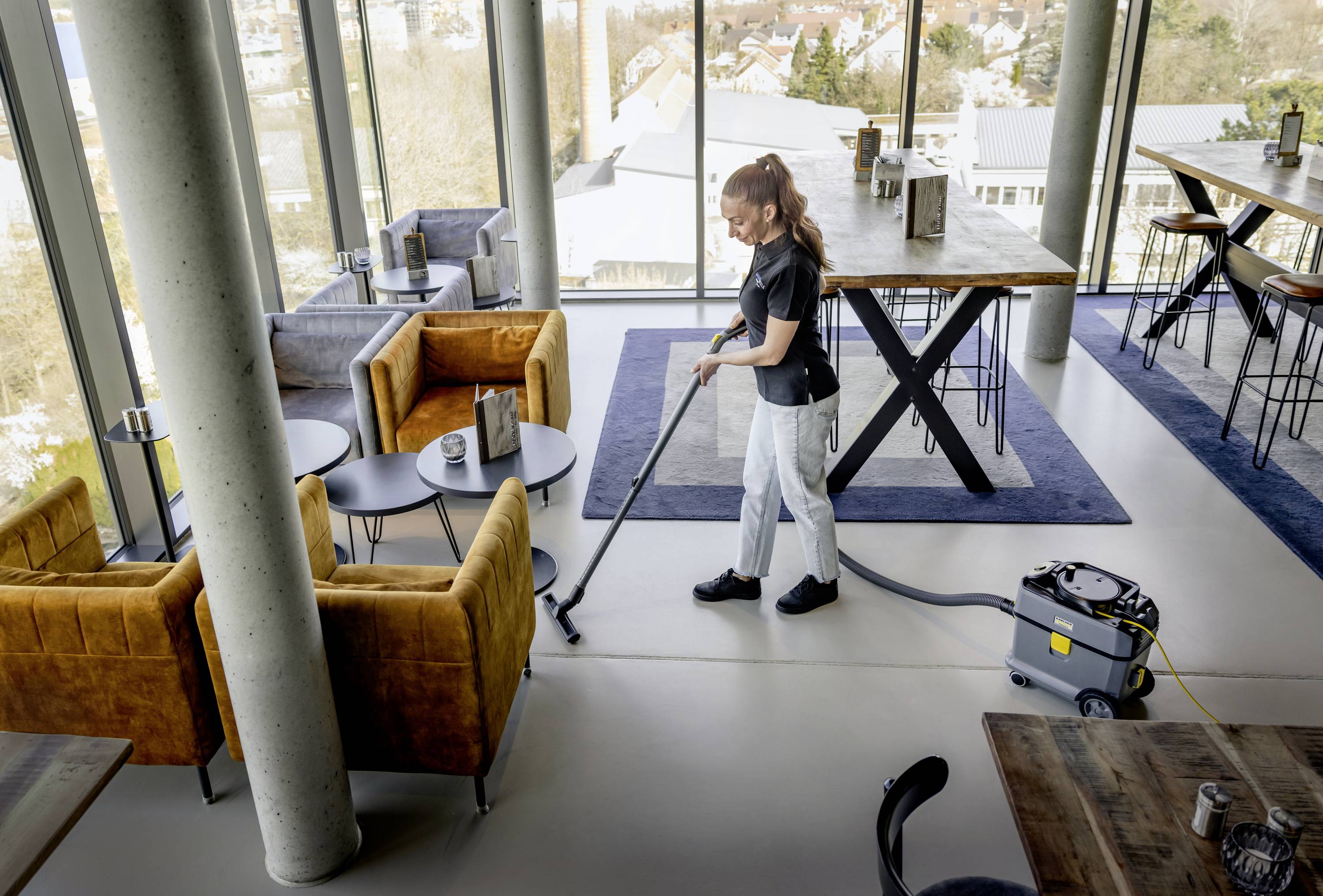 En person dammar golvet i ett modernt, väl belyst café med stora fönster. Bekväma sittplatser syns.