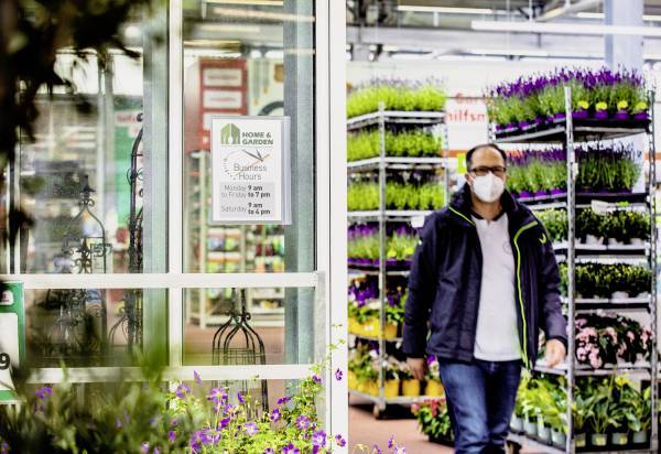 En person med munskydd står i ett trädgårdscenter bredvid blommande växtställ. En skylt visar öppettiderna.