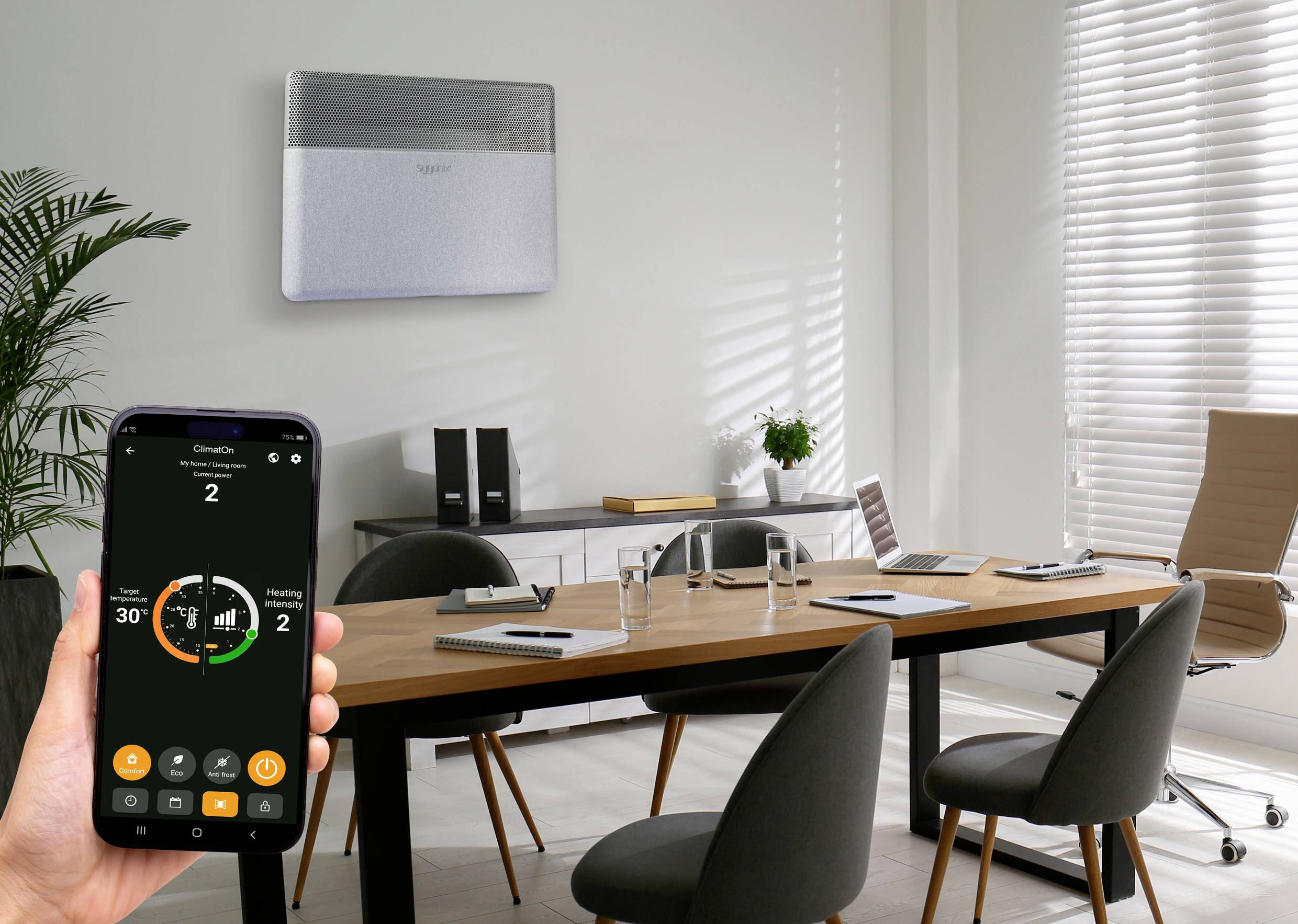 Kontor med bord och stolar, i bakgrunden en värmeanordning på väggen. En hand håller en smartphone med en öppen värmestyrnings-app.