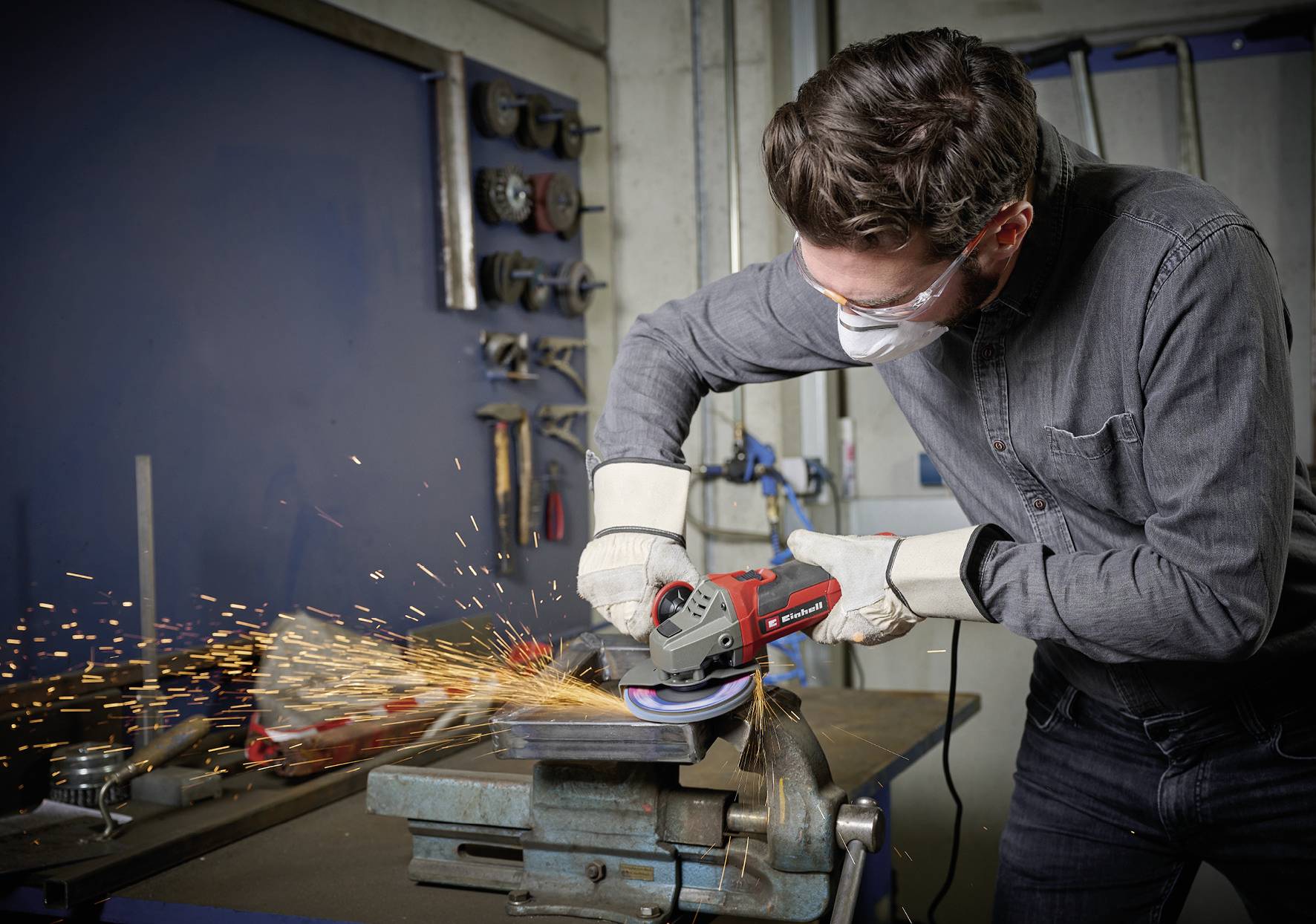 En man sliper metall med en slipmaskin i en verkstad. Han bär skyddsglasögon, mask och handskar. Gnistor flyger.