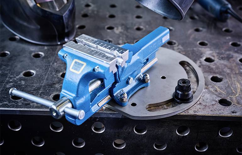 En blå skruvstäd är monterad på en metallarbetsplats. I bakgrunden syns verktyg och en svetshjälm.