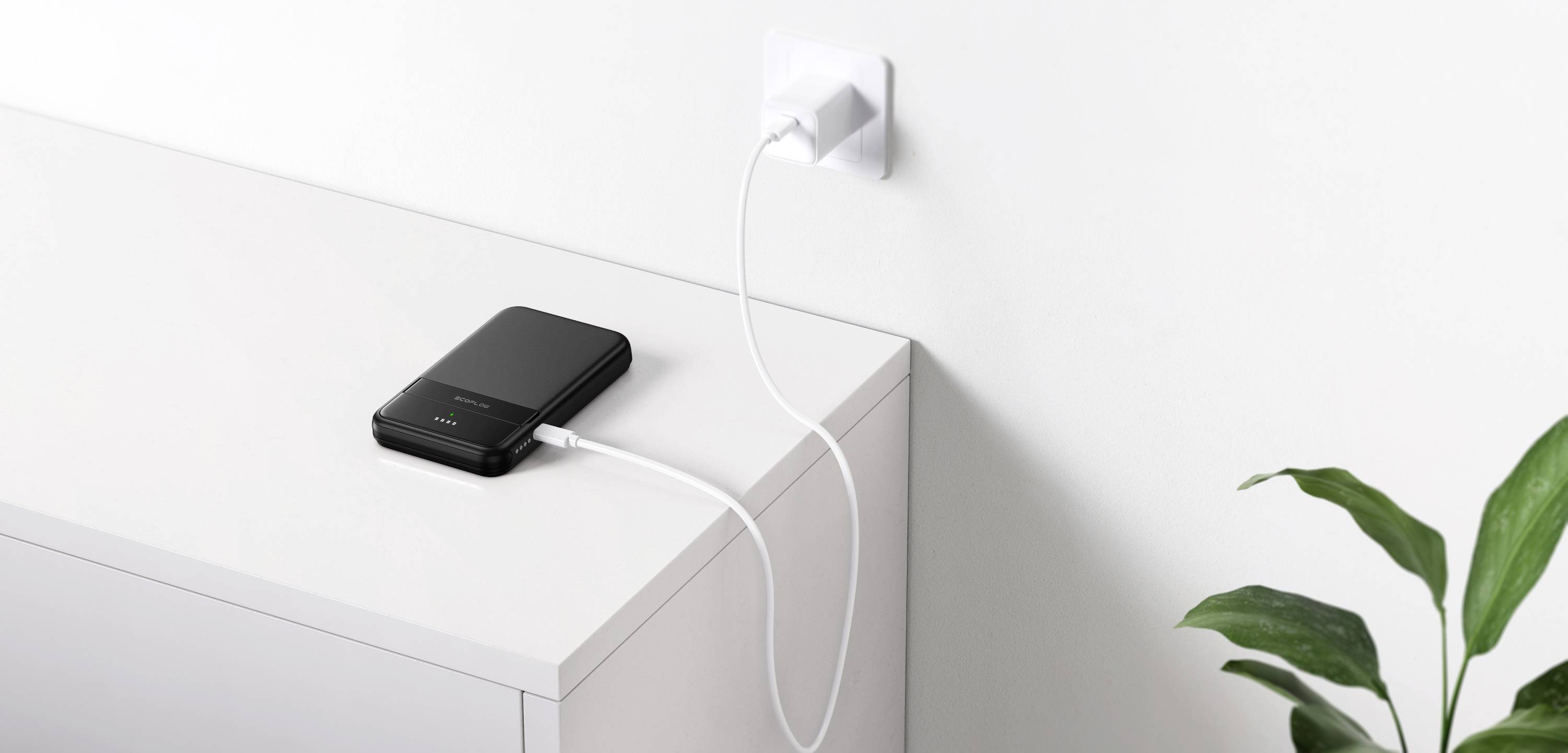 En svart batteridriven enhet laddas med en kabel från ett vägguttag på ett vitt skåp. Bredvid står en grön växt.