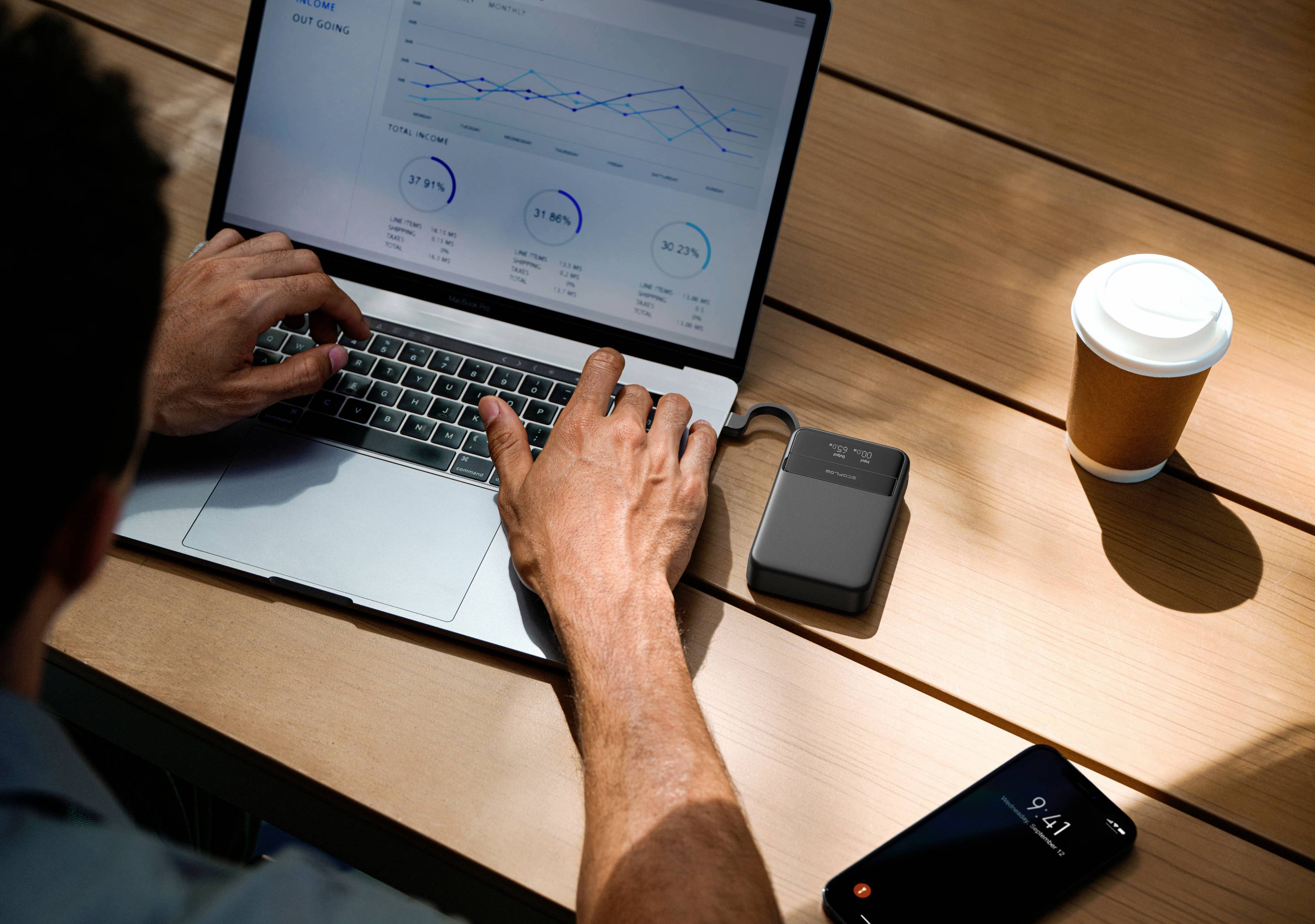 En person arbetar på en bärbar dator med ett diagram på skärmen; bredvid ligger en smartphone, en kaffekopp och en extern powerbank på ett träbord.