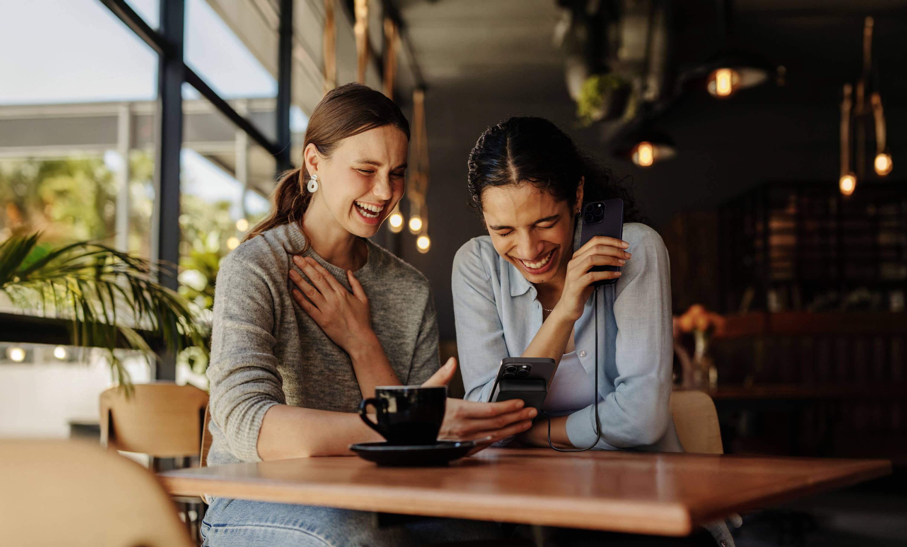 Två skrattande kvinnor sitter på ett café, tittar tillsammans på en smartphone och verkar glada och avslappnade.