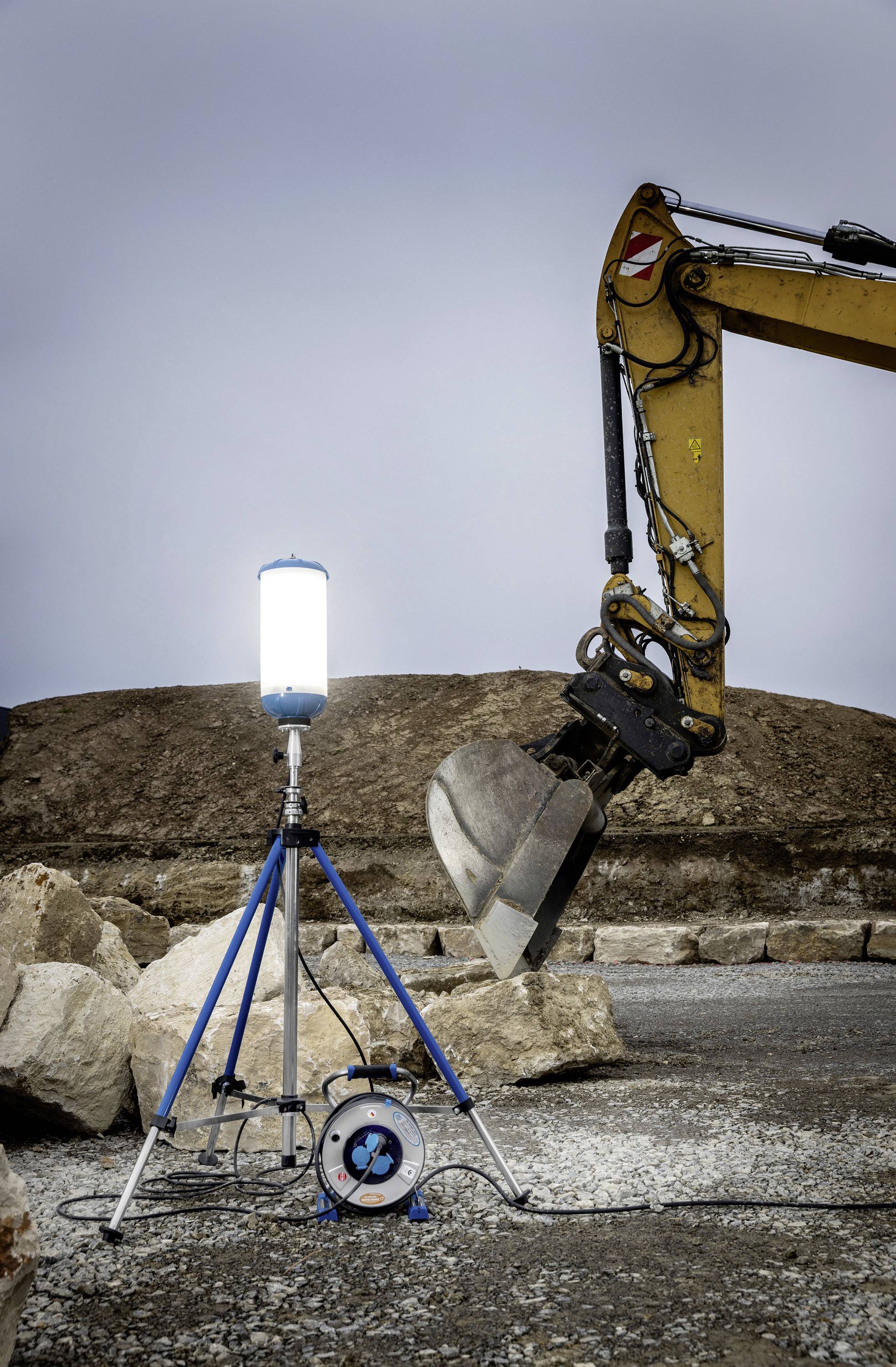 En byggarbetsplats visar en upplyst strålkastare på ett stativ bredvid en grävmaskins arm med skopa, omgiven av stenar.
