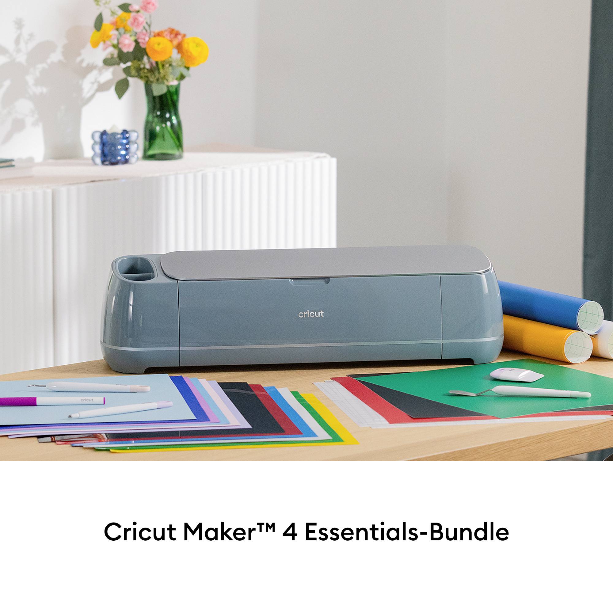 En Cricut Maker™ 4 står på ett bord med färgglada pappersark och verktyg. I bakgrunden en vas med blommor.