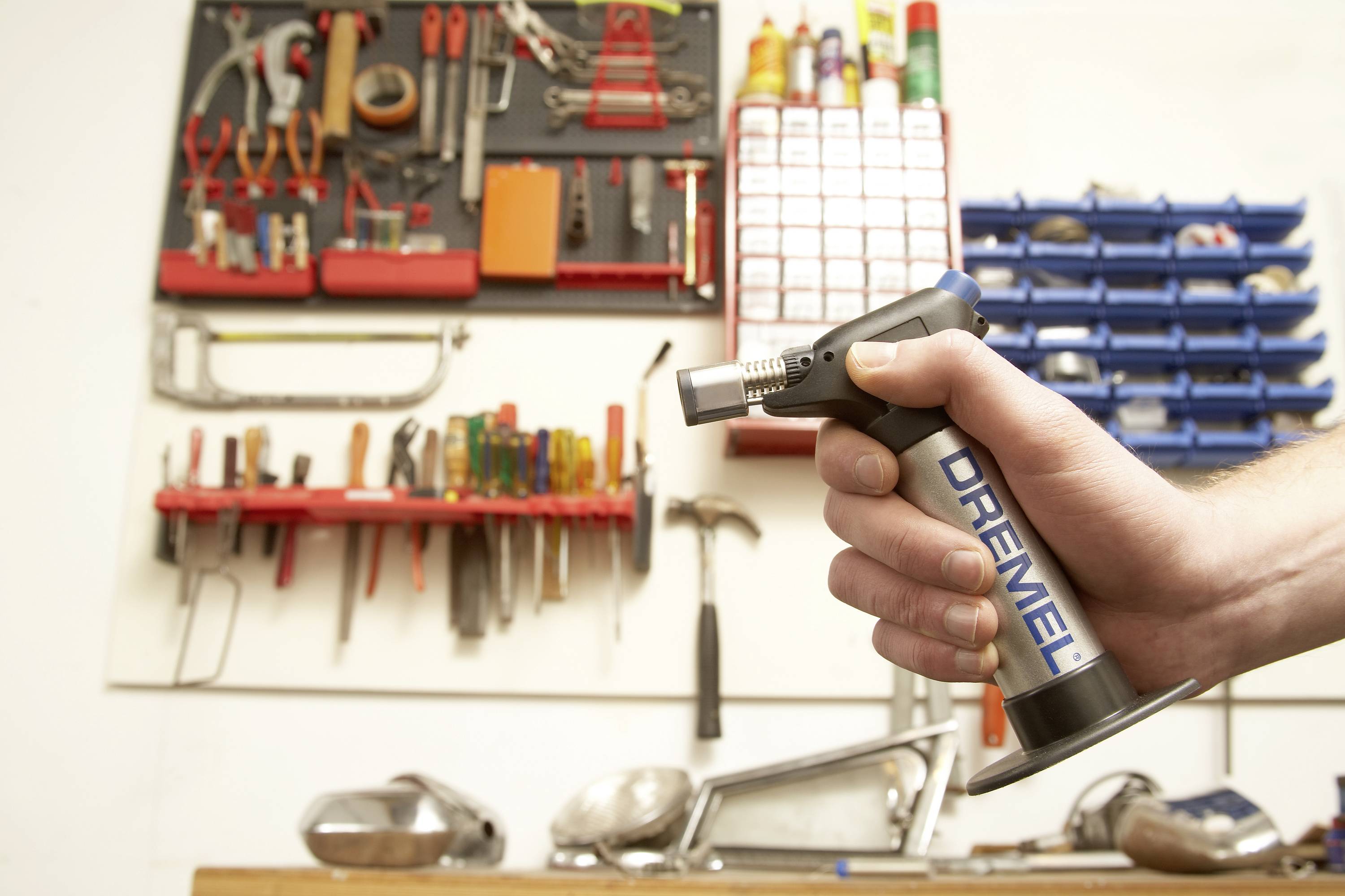 En hand håller en Dremel-lödkolv i en verkstad med krokar på väggen där verktyg och material är organiserade.