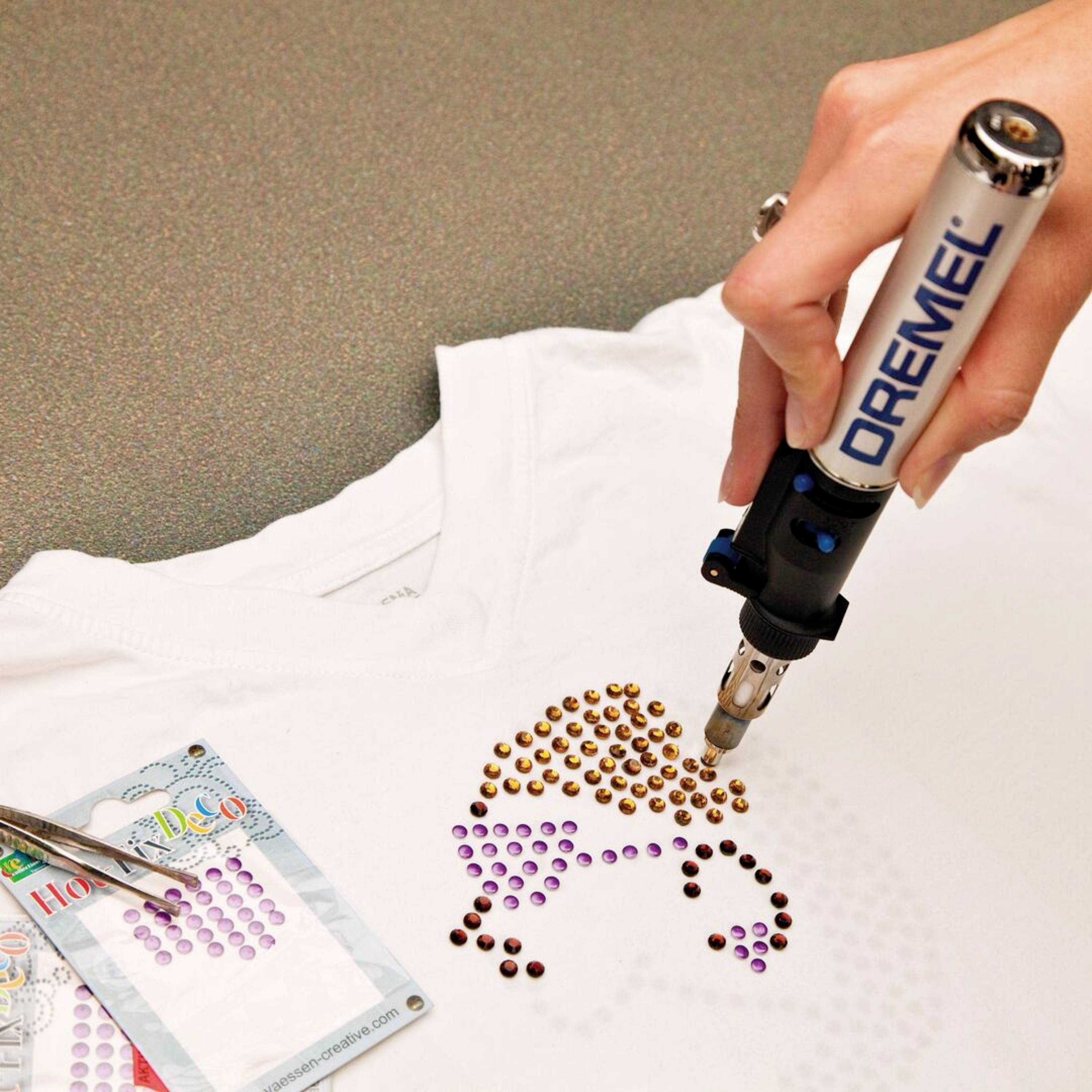 En hand använder ett verktyg för att fästa ett mönster av färgglada nitar på en vit t-shirt. Till höger syns ett Dremel-verktyg.