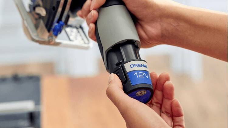En person håller och för in ett blått och grått 'Dremel 12V' batteri i ett verktyg, med fokus på batteritillsättningsprocessen.