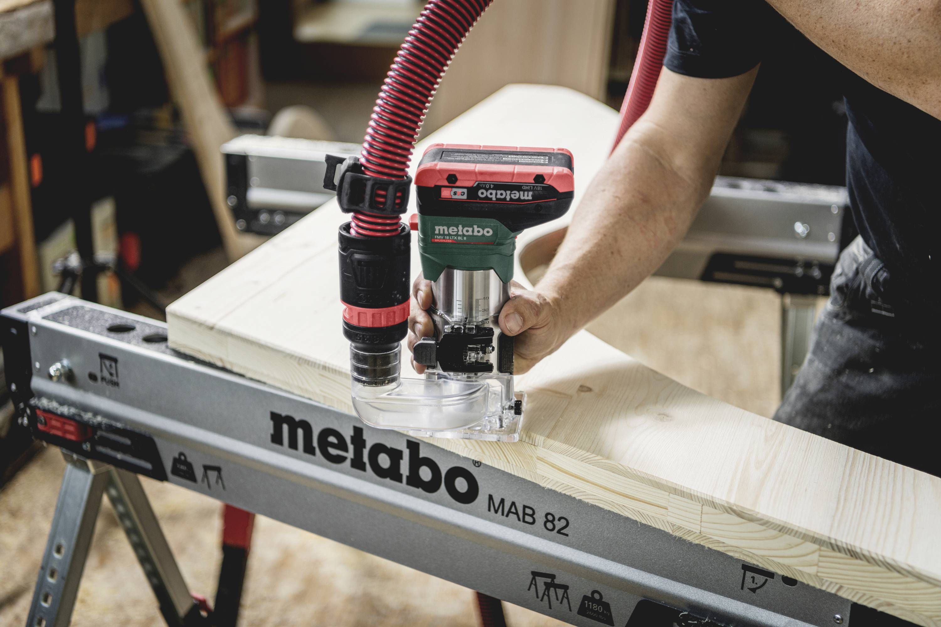 En person arbetar med en handhållen fräs på ett ljust trästycke, där fräsen är fastsatt på ett Metabo-verktygsstånd.