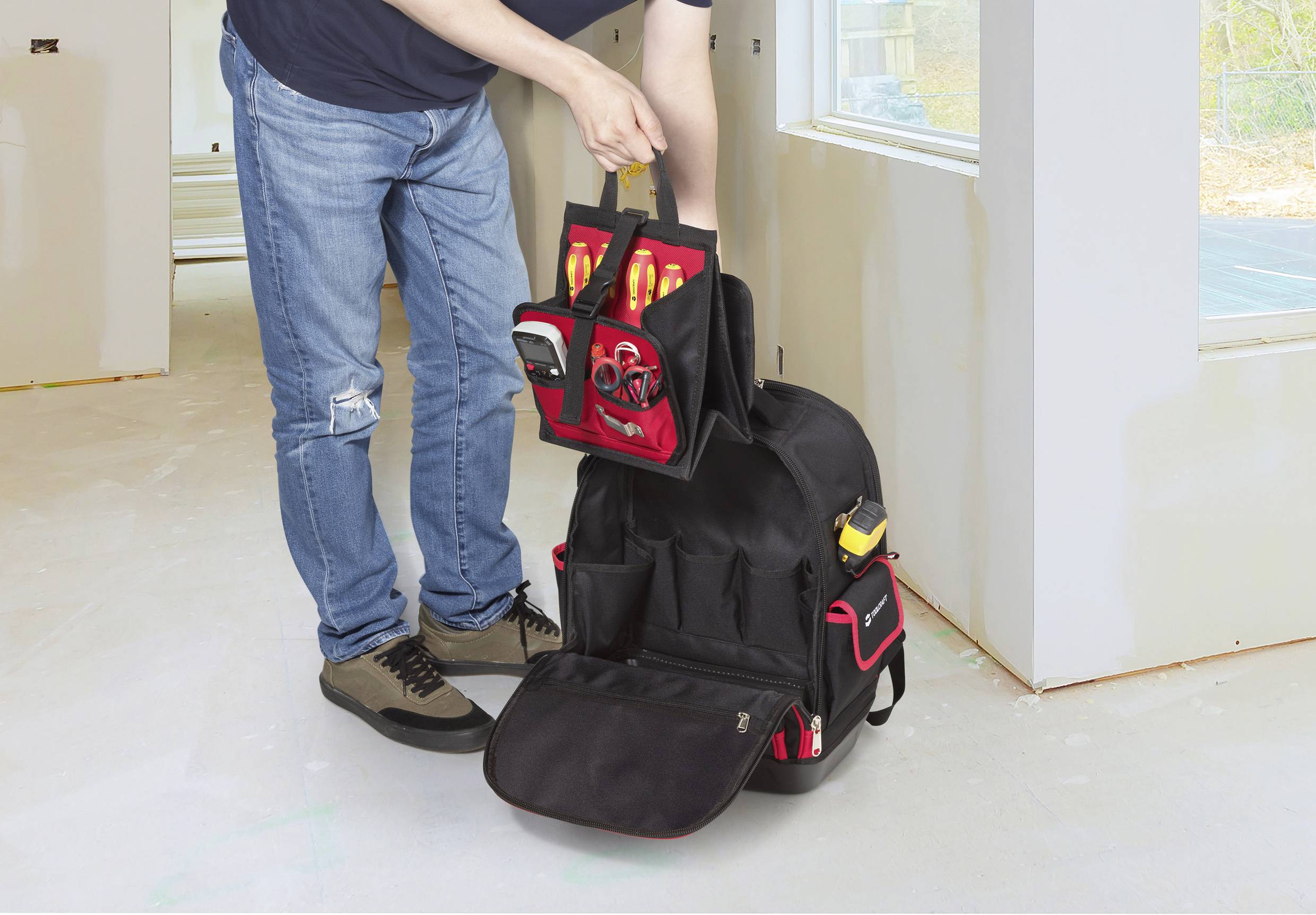 En person som organiserar verktyg i en svart ryggsäck med röda detaljer, inuti ett väl upplyst rum under renovering.
