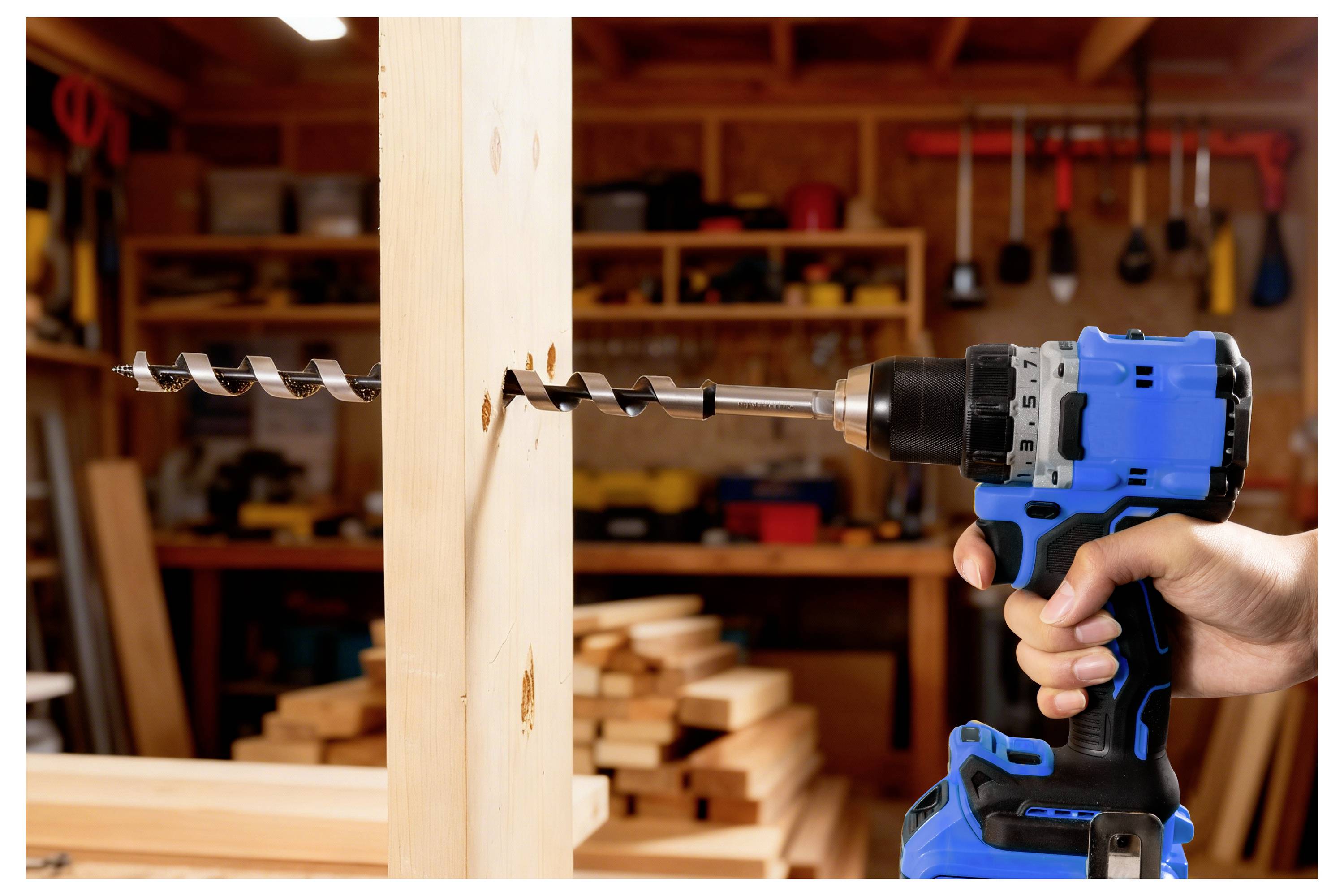 En person använder en blå sladdlös borrmaskin med ett stort borr för att göra hål i en träbalk inuti en verkstad med verktyg i bakgrunden.