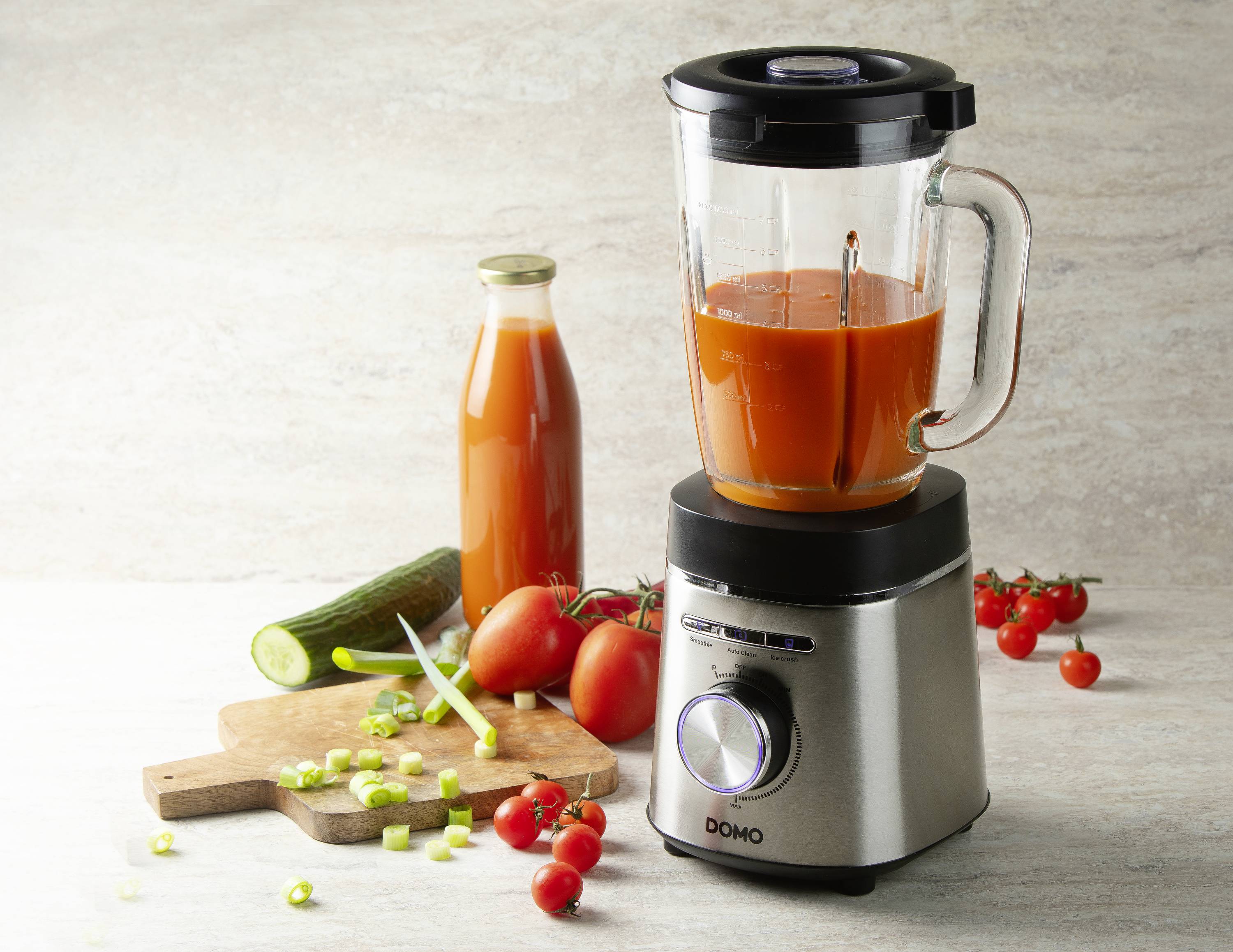 En mixer fylld med röd vätska står bredvid en juiceflaska, tomater och gurkor på en skärbräda.