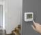 En hand justerar en digital termostat som visar '24.5°C'. Den är monterad på en grå vägg intill en trätrappa i ett modernt hem.