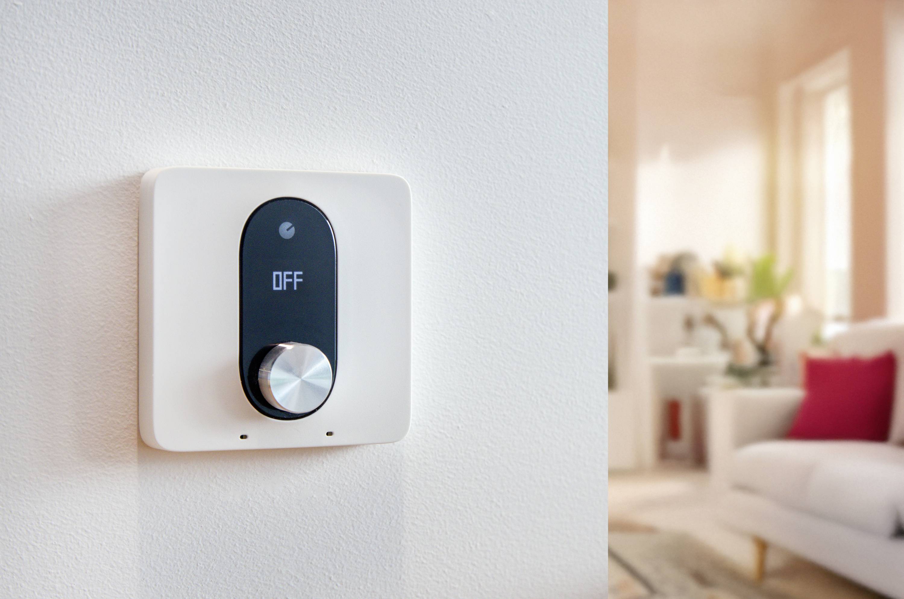 En digital termostat på en vägg visar 'AV'. I bakgrunden syns ett suddig inrett vardagsrum med soffa och kuddar.