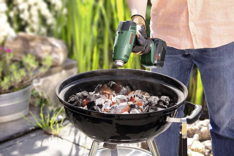 En person tänder kol på en grill med en heteluftspistol. Trädgårdsmiljö i bakgrunden.