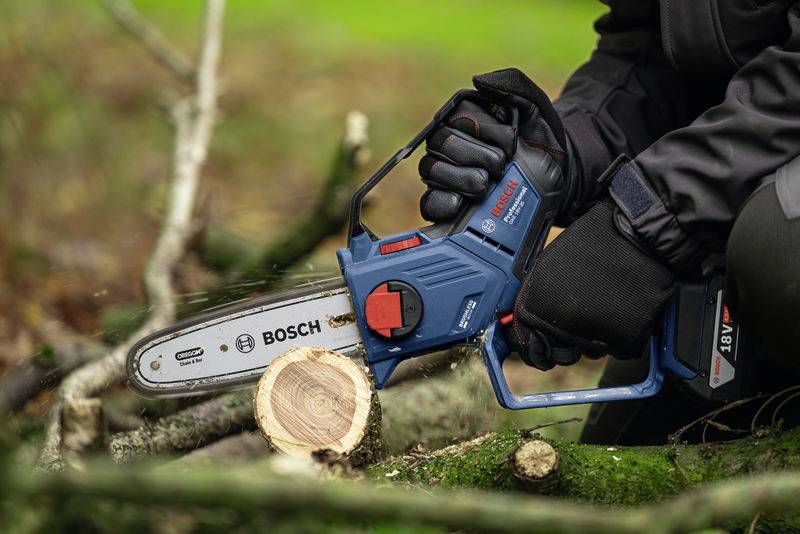 En person sågar en trästock med en blå Bosch-sladdlös kedjesåg på en grön äng.