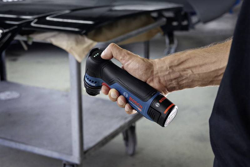 En hand håller en Bosch-batteridriven skruvdragare som är förberedd för arbete på en mörk yta, med en oskarp verkstad i bakgrunden.