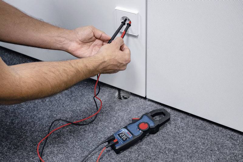 En person mäter spänningen i ett eluttag med ett multimeter. Displayen visar '225 V'.