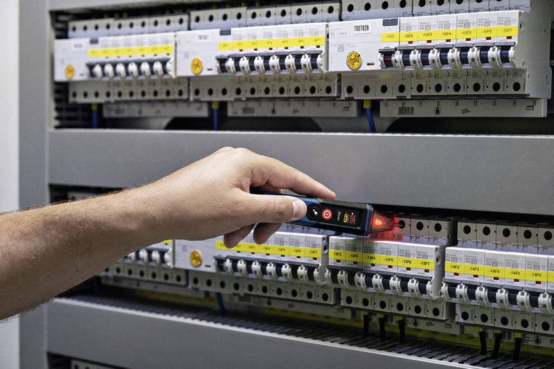 En hand håller ett spänningstestinstrument framför en elektrisk säkringslåda för att kontrollera spänningen.