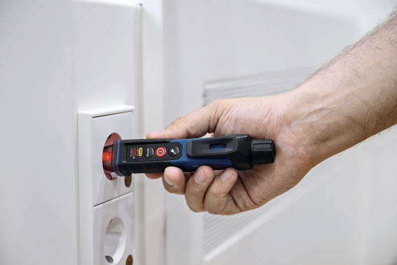 En hand håller ett spänningstestinstrument mot ett eluttag för att kontrollera elektrisk spänning.