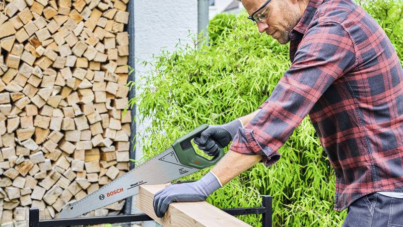 En man i arbetsklädsel sågar ett träbräde utomhus med en handsåg. I bakgrunden syns staplade träbitar och gröna växter.