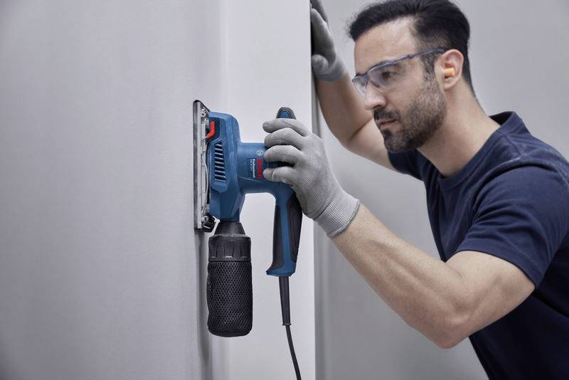 En man sliper en vägg med en elektrisk slipmaskin. Han bär skyddsglasögon och handskar och arbetar fokuserat.