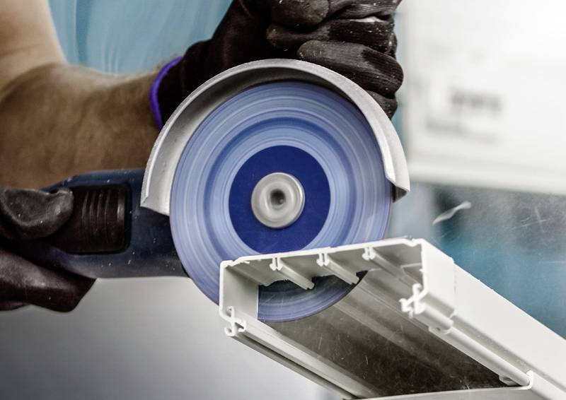 En person sågar ett metallstycke med en blå cirkelsåg. Skyddsutrustning är synlig, med fokus på precision under sågningen.